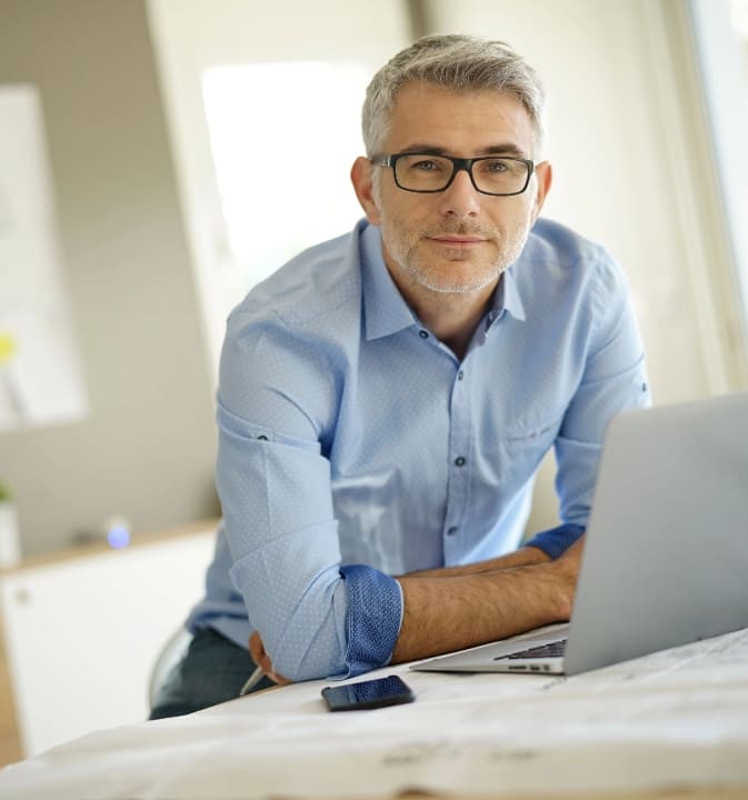 Man wearing blue shirt and glasses resting his arms on table with open laptop in front of him
